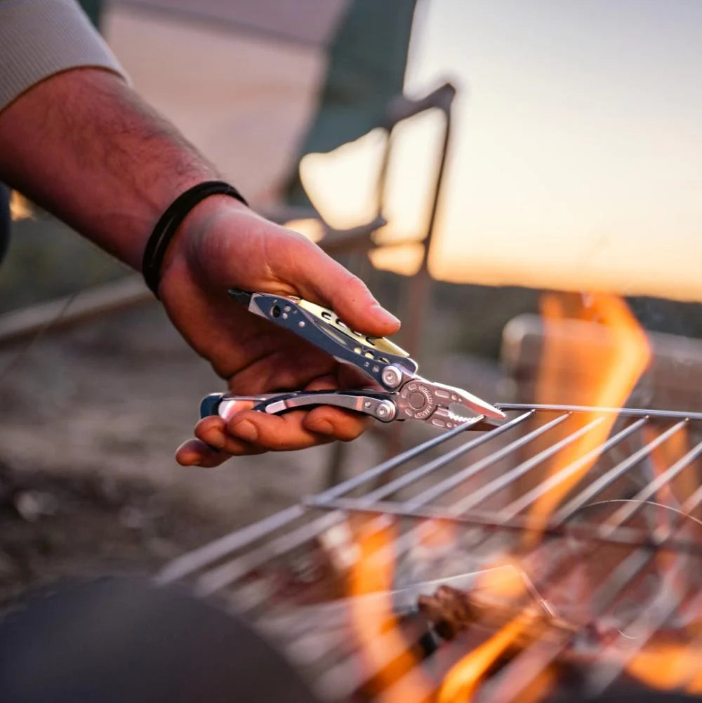 Hand holding a multi-tool over a campfire with lanterns in the background