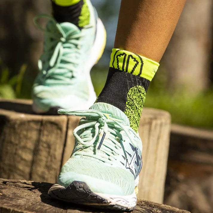 Person wearing green running shoes and black socks with yellow accents on a wooden log outdoors.
